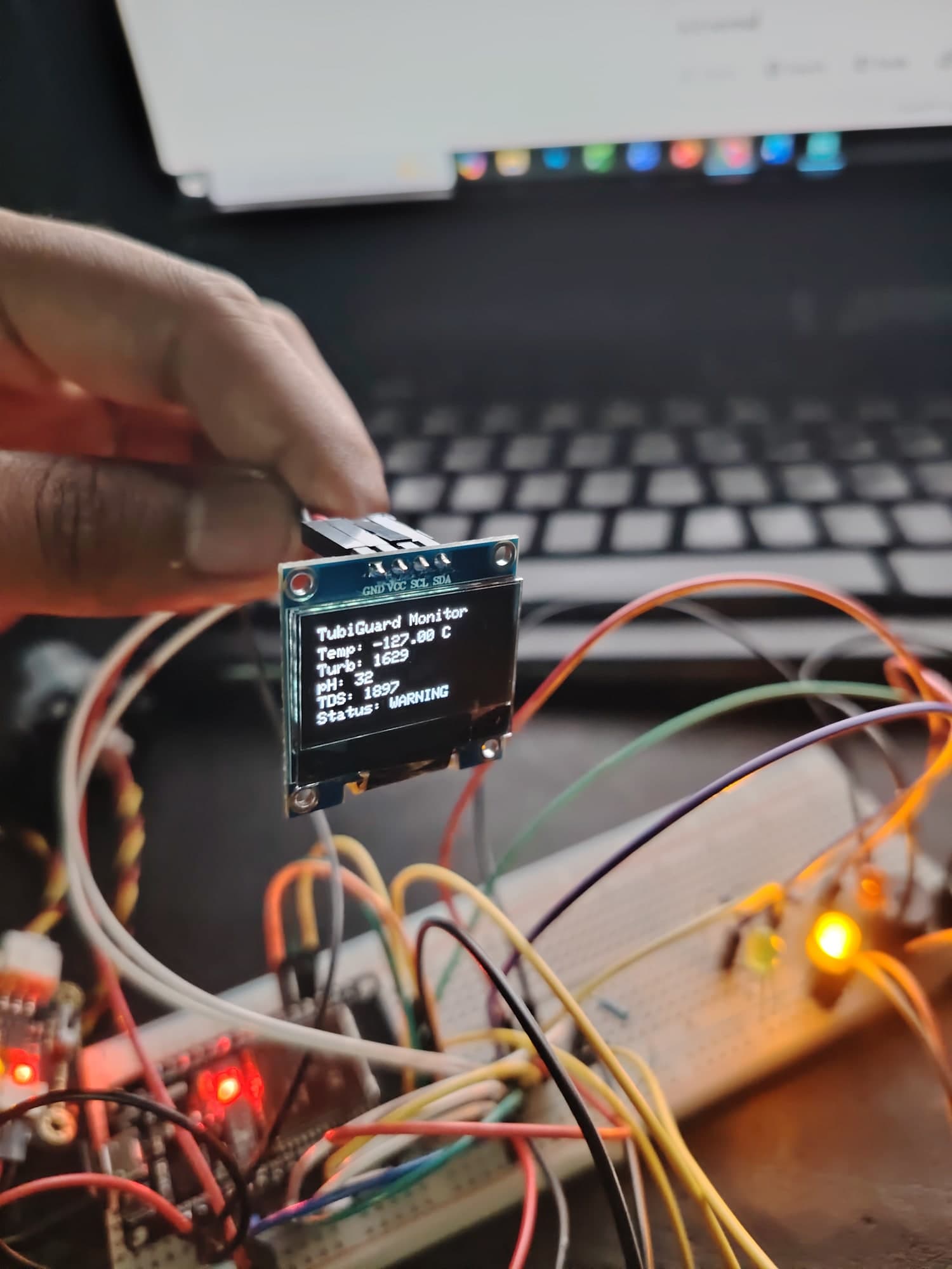 The TubiGuard display held in hand during bench testing, with the breadboard build behind it.